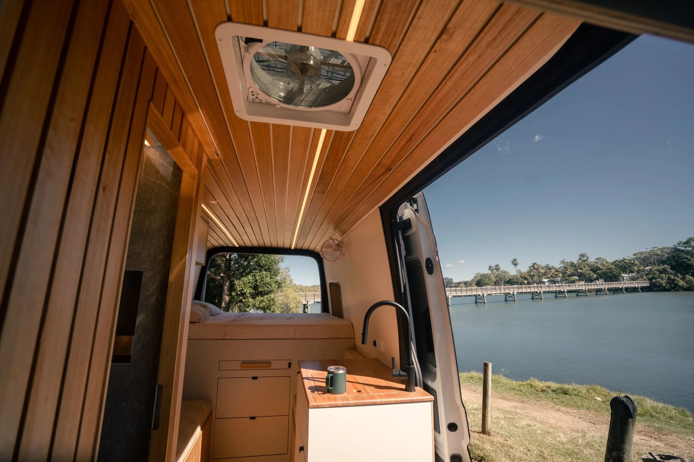 Morning light inside the campervan, a coffee cup on the bench and a jetty across still water