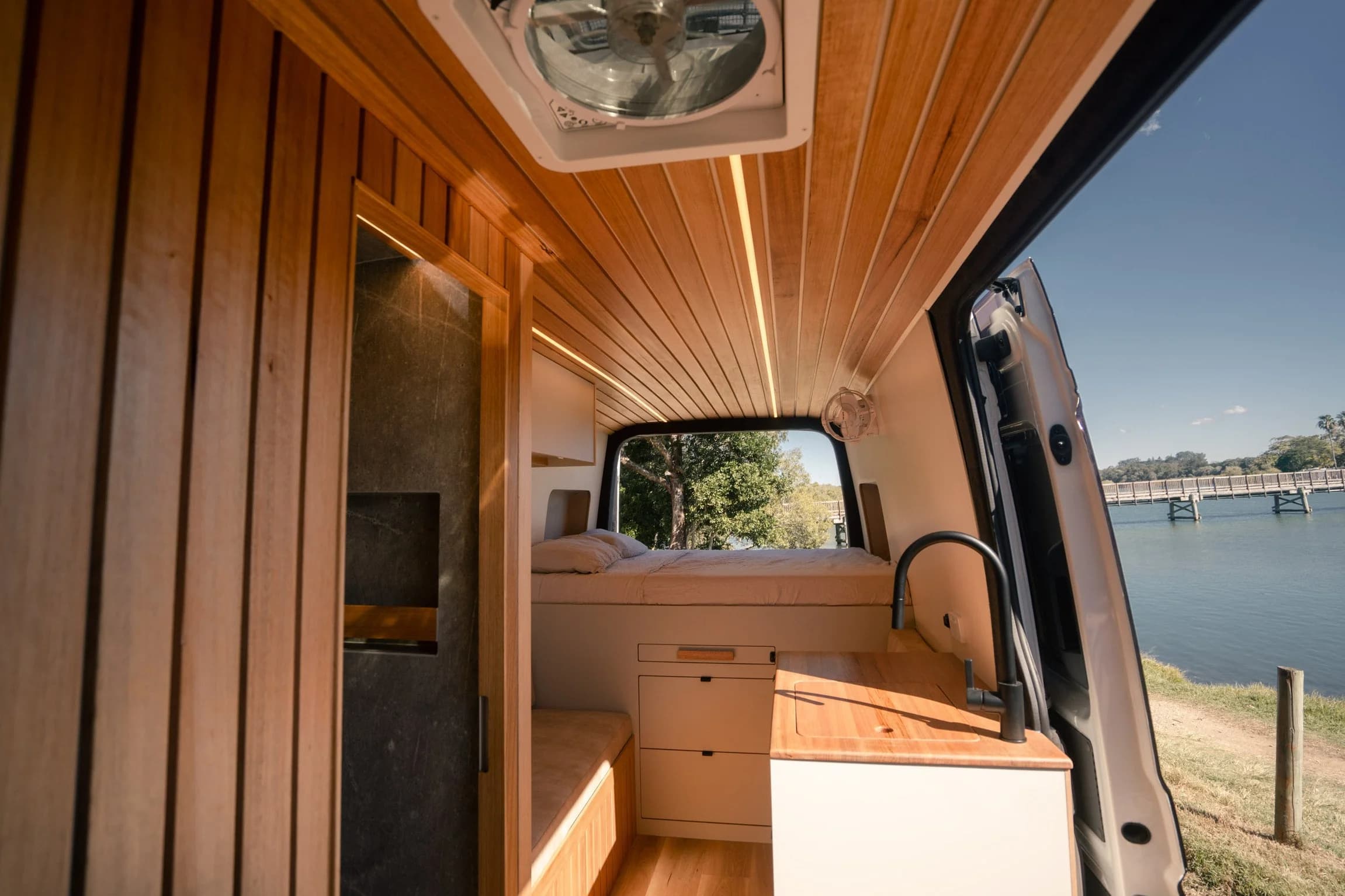 Interior of the Bondi Van Co campervan with timber ceiling, bed and galley, side door open onto water