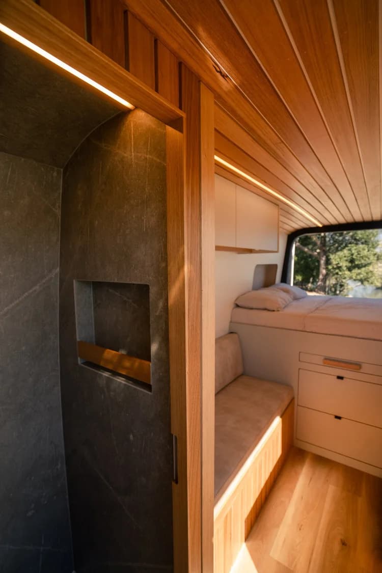 Open shower door revealing stone walls, with the bed and bench seating beyond